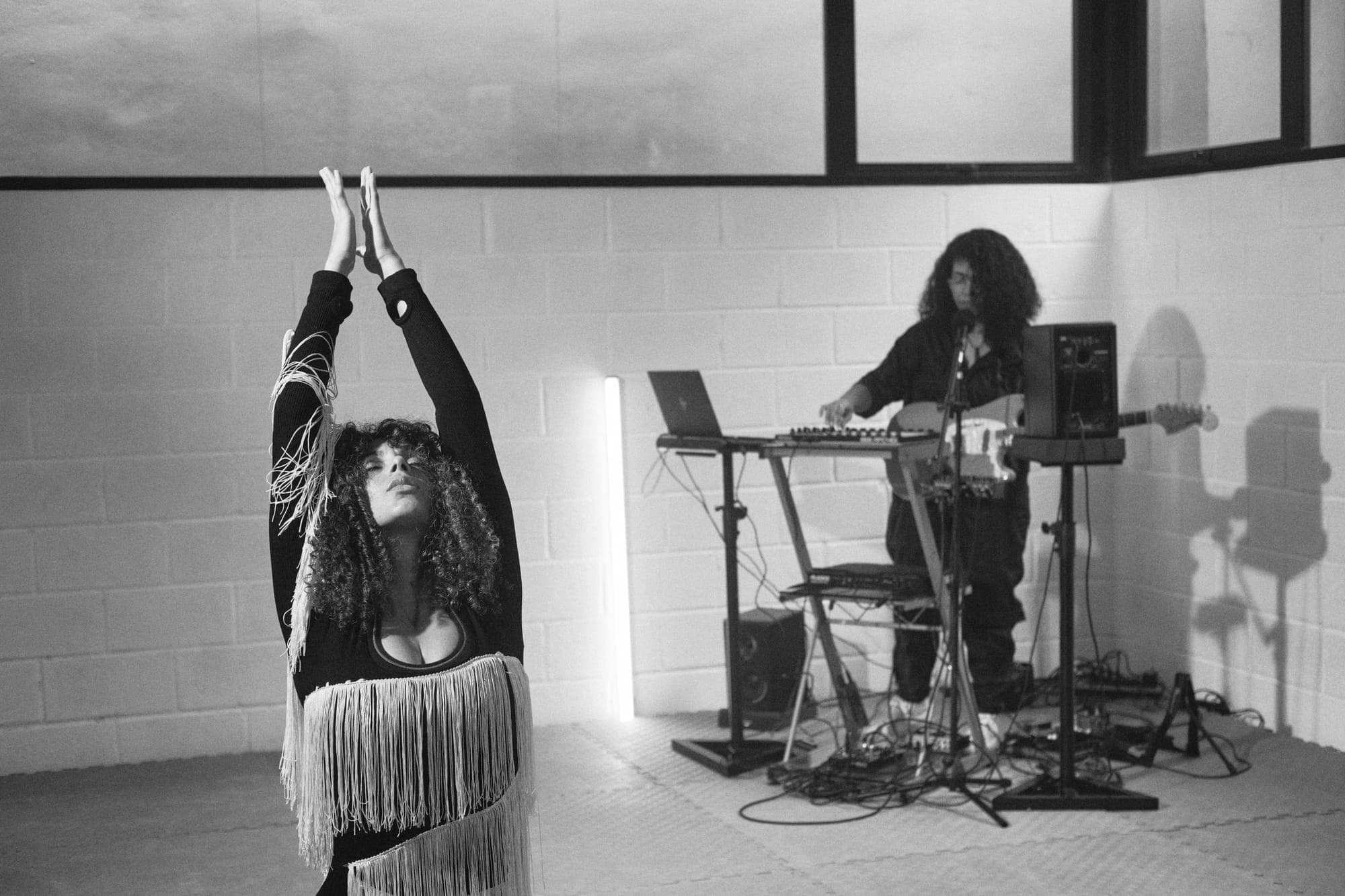 La bailarina alza los brazos con el top de flecos mientras Maria Lunares toca al fondo, junto al soporte del teclado.