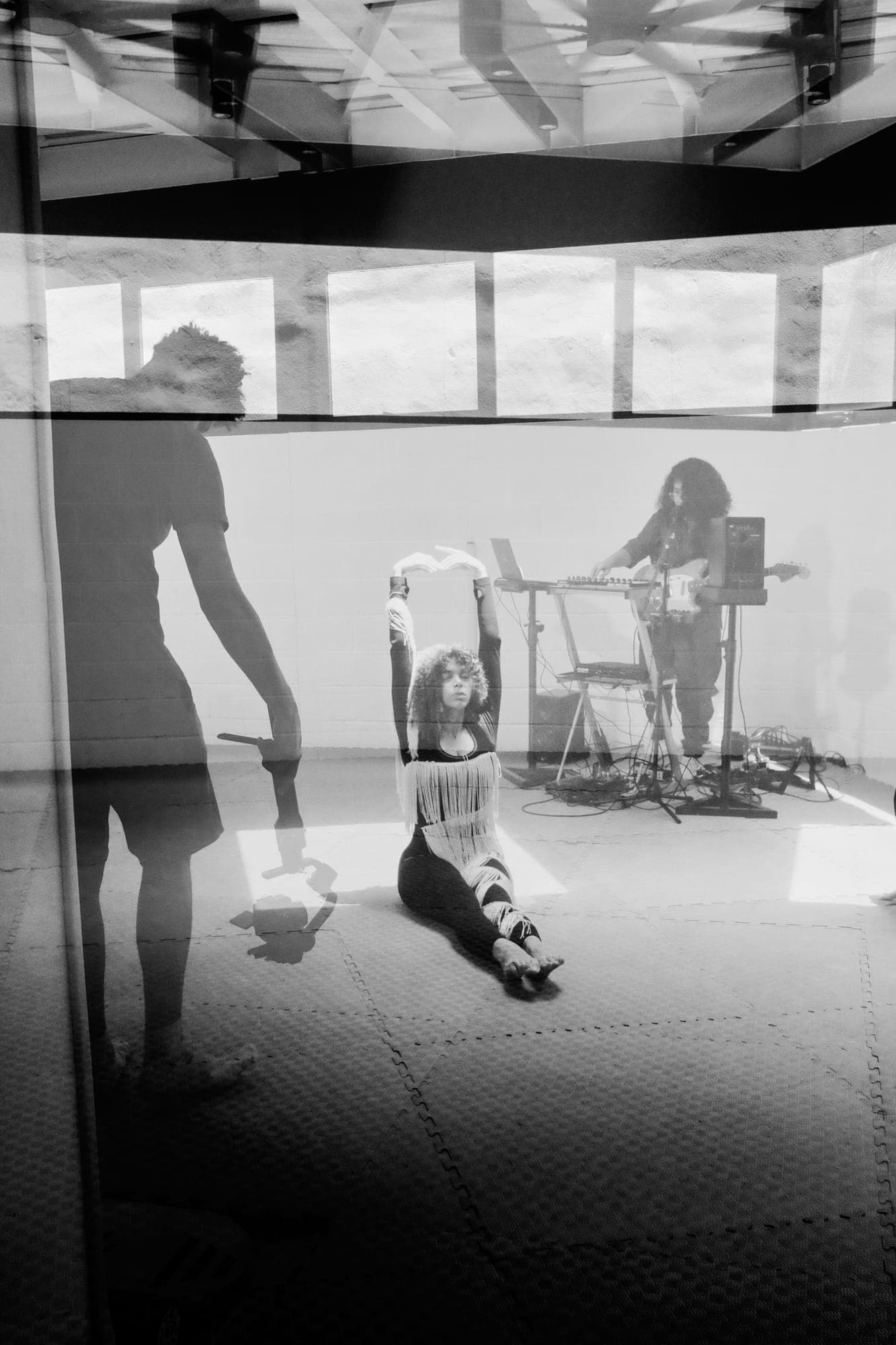 Doble exposición de la sala: la bailarina sentada en el suelo con los brazos en alto y Maria Lunares tocando al fondo.