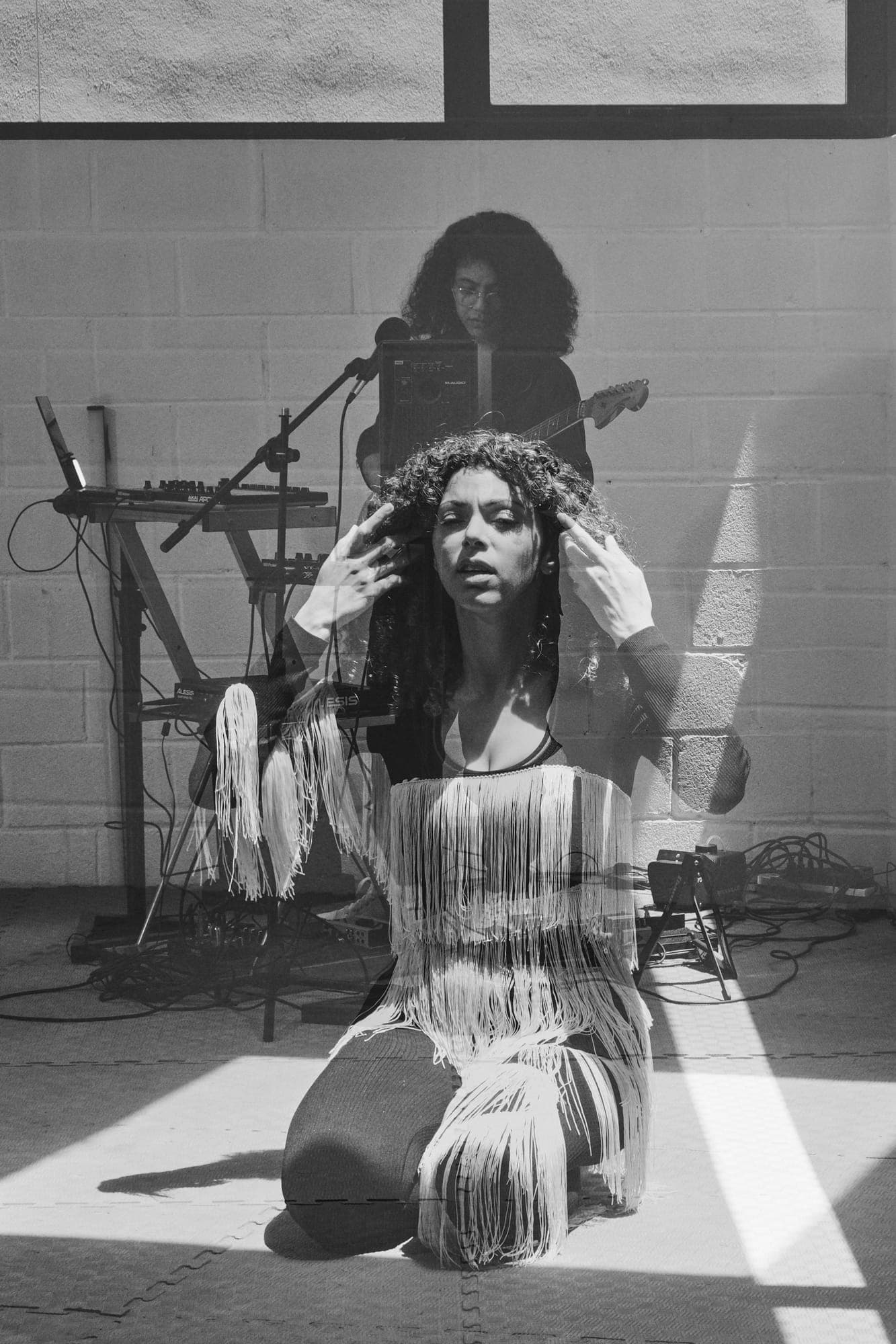 Doble exposición: la bailarina de rodillas en un haz de luz con las manos en la cabeza y Maria Lunares detrás, al micrófono.