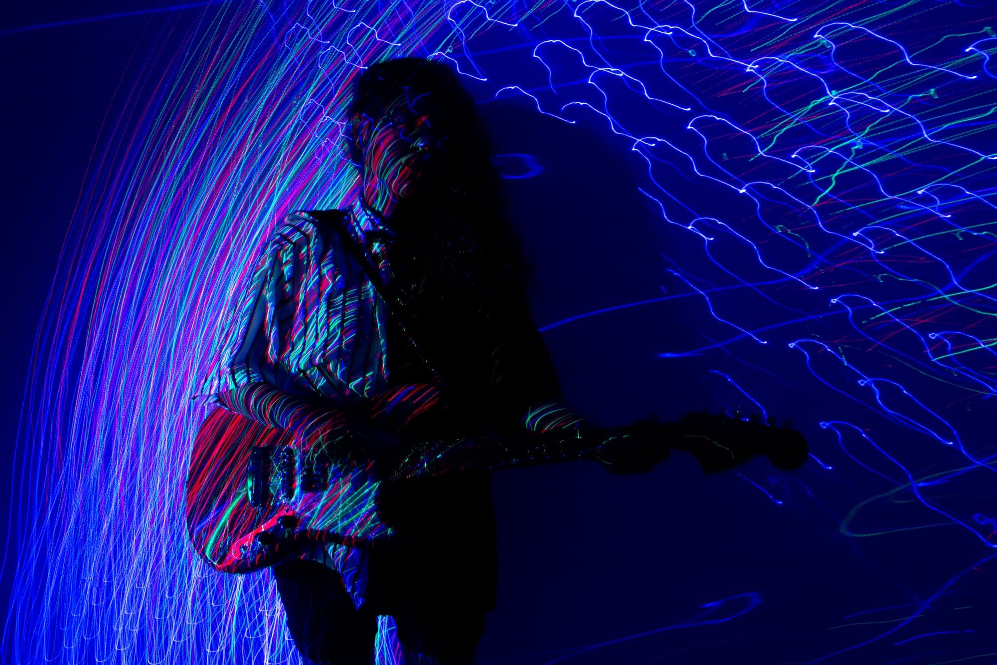 Tocando la guitarra entre trazos de luz azul y magenta que cruzan el encuadre en exposición larga.