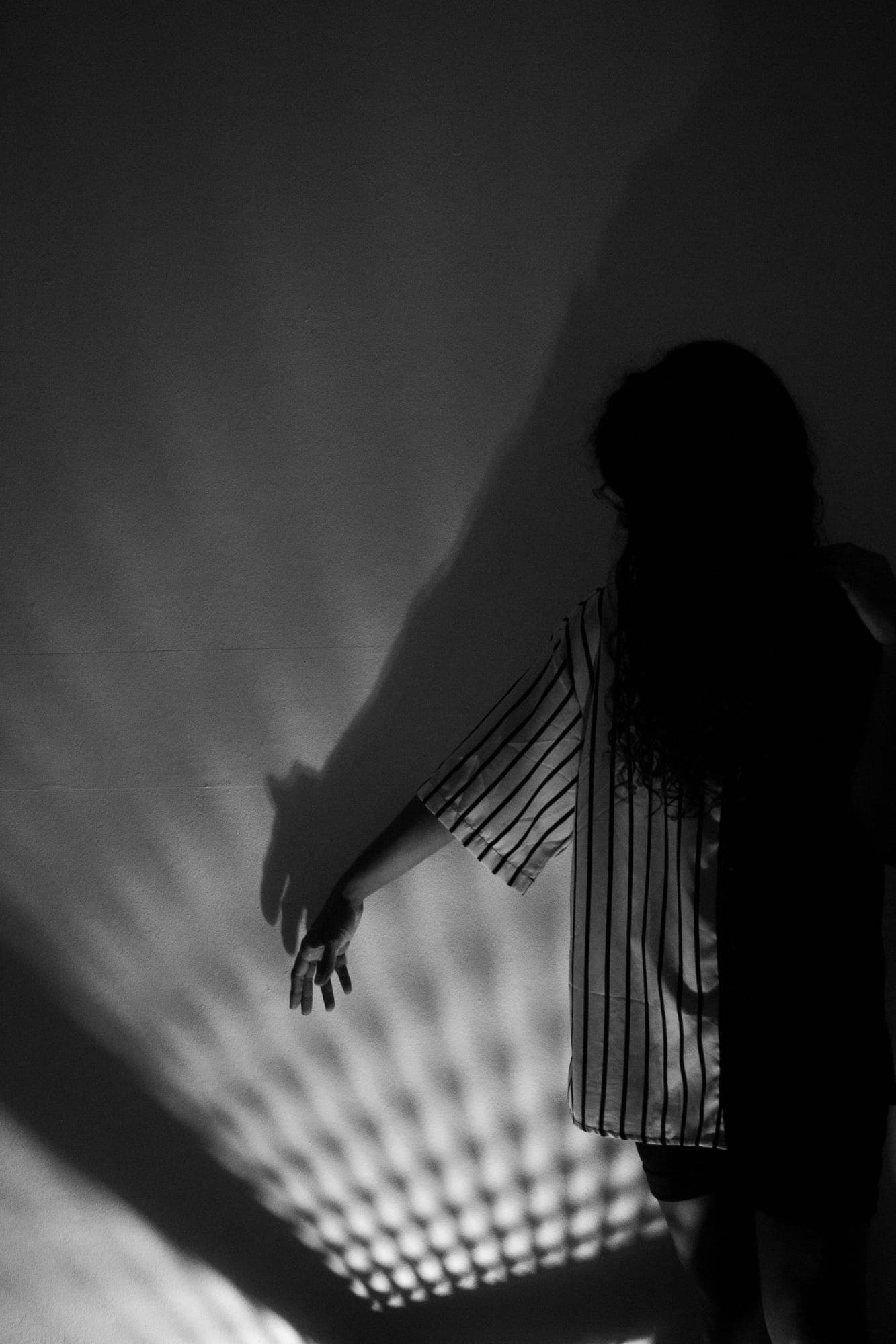 Blanco y negro: camisa de rayas contra una pared, el brazo extendido y una sombra de celosía proyectada a su lado.
