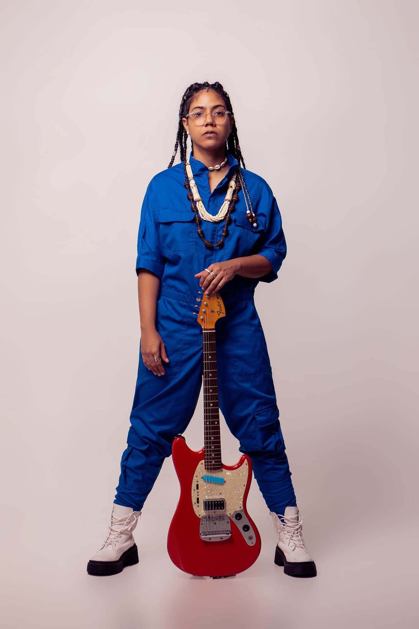 Retrato de cuerpo entero con mono de trabajo azul y botas blancas, sosteniendo de pie una guitarra Fender Mustang roja.