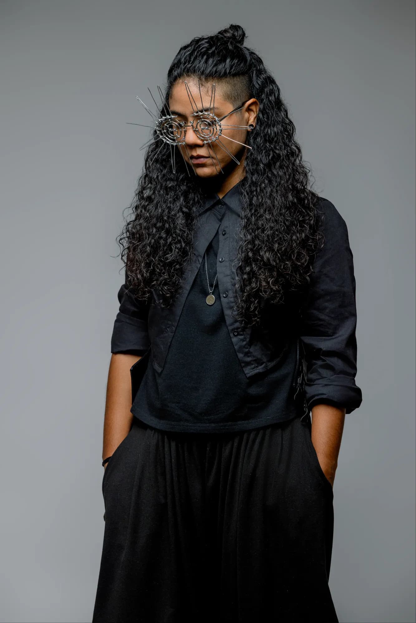 Camisa negra abierta sobre camiseta, medio moño y las gafas de púas, con la cabeza inclinada y las manos en los bolsillos.