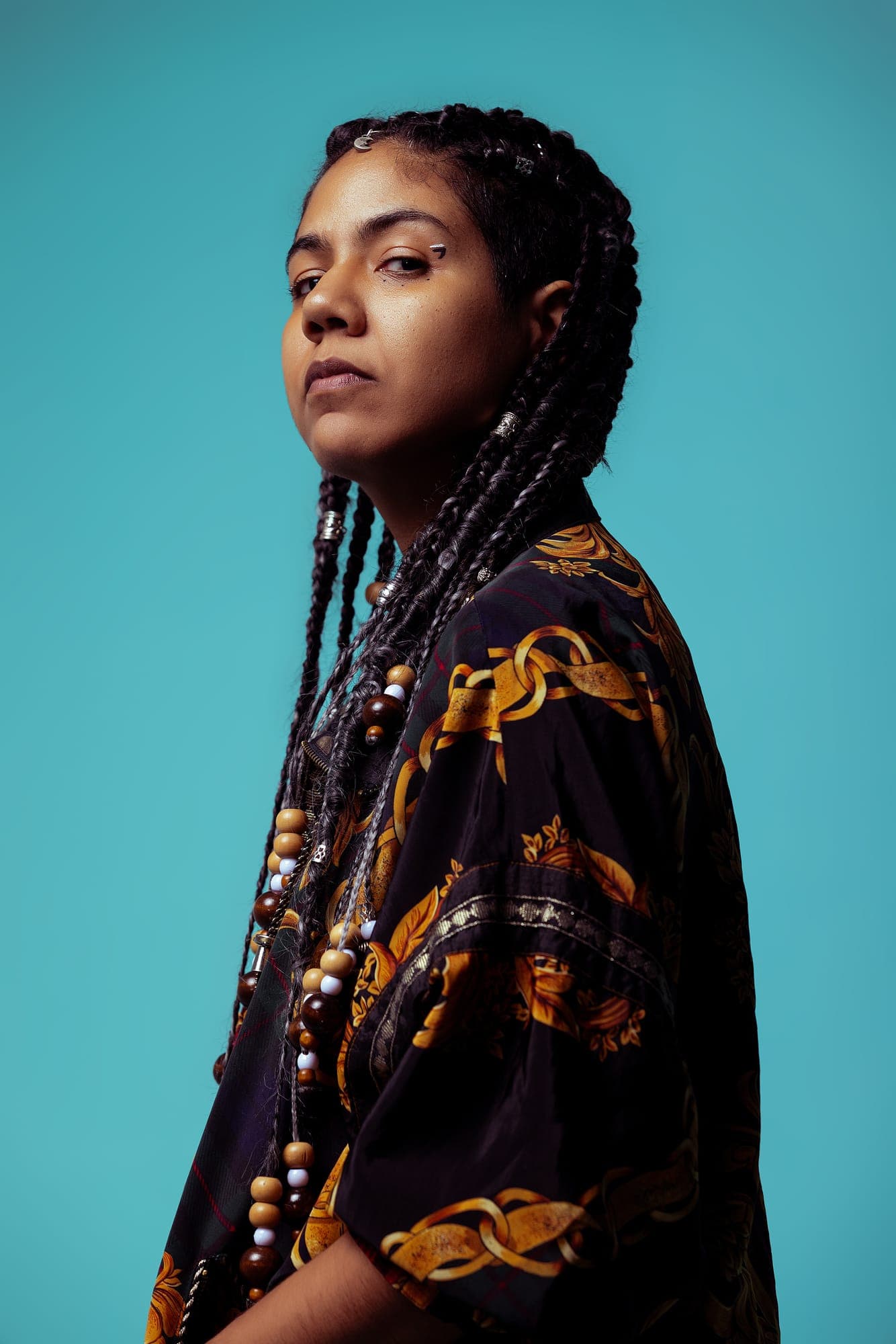 Maria Lunares de perfil sobre fondo turquesa, con trenzas largas rematadas en cuentas de madera y una chaqueta oscura de estampado dorado.