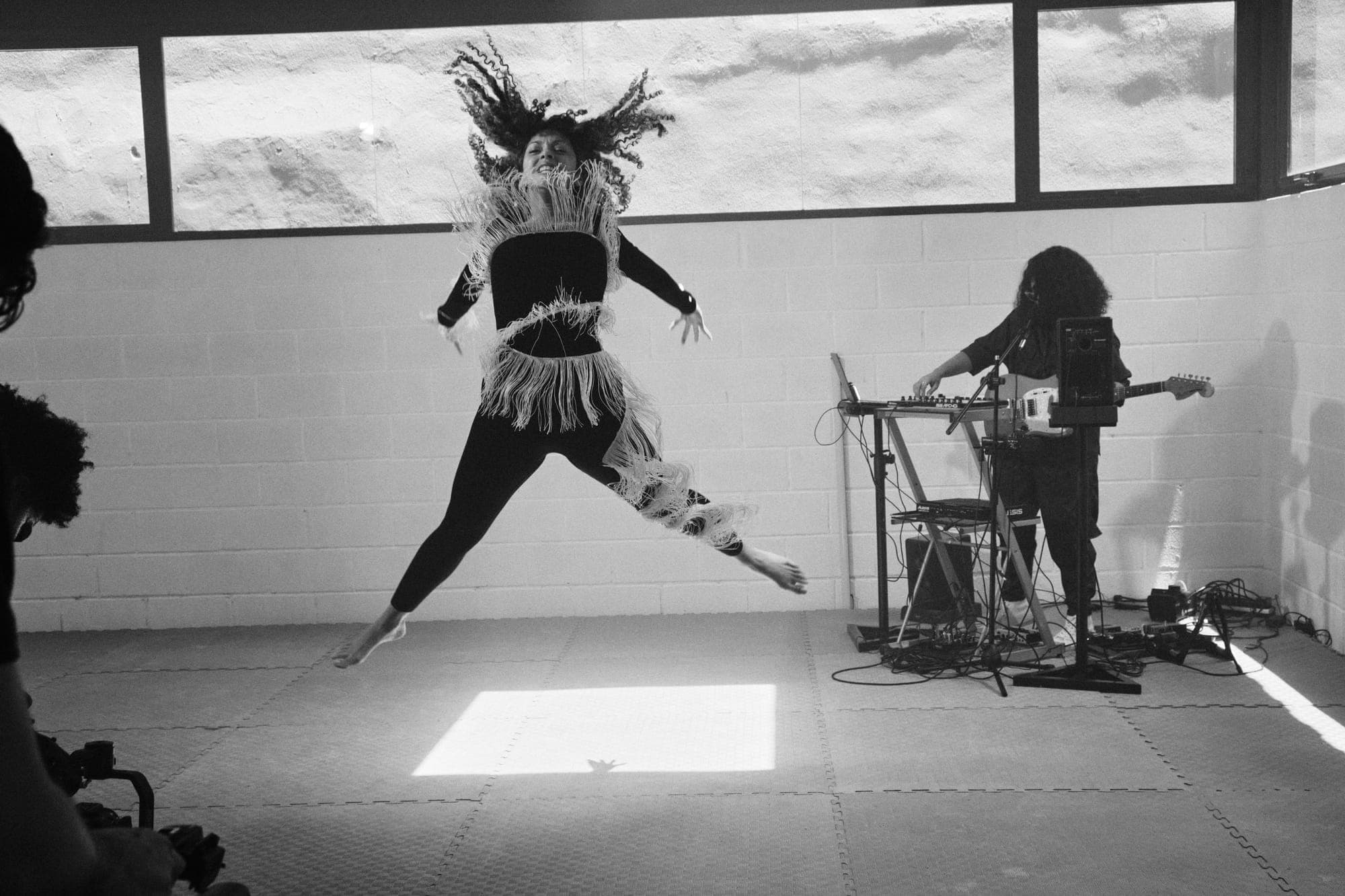 Una bailarina salta descalza con un traje de flecos mientras Maria Lunares toca la guitarra junto a la pared del ensayo.